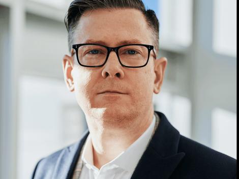 A professional portrait of a man wearing glasses, a blue blazer, and a white shirt standing in an office environment.